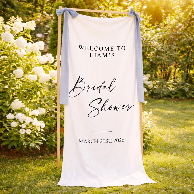 May include: A white fabric banner with the text "WELCOME TO LIAM'S Bridal Shower MARCH 21ST, 2026" in black script. The banner is suspended from a wooden frame with light blue ribbons. The background features greenery and sunlight.