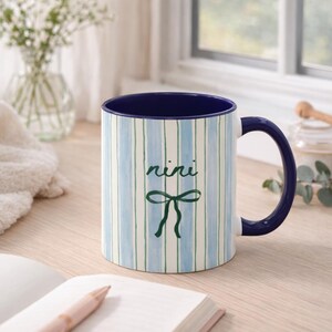May include: A ceramic mug with a blue interior and handle. The exterior features vertical stripes in blue and green, with the word "nini" and a bow design. The mug is on a wooden surface.