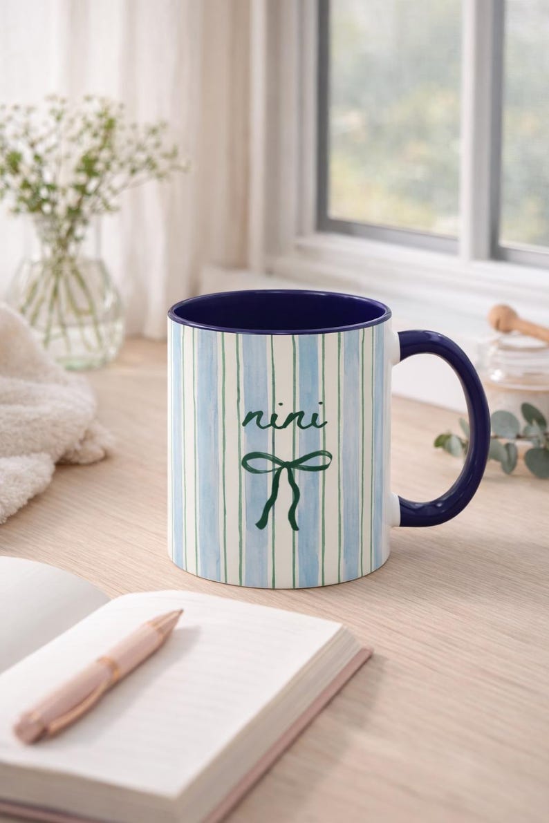 May include: A ceramic mug with a blue interior and handle. The exterior features vertical stripes in blue and green, with the word "nini" and a bow design. The mug is on a wooden surface.