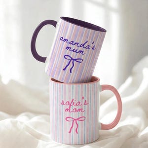 May include: Two ceramic mugs stacked on top of each other. The top mug has a purple handle and the text "amanda's mum" in cursive, with a bow illustration. The bottom mug has a pink handle and the text "sofia's mom" with a bow illustration.