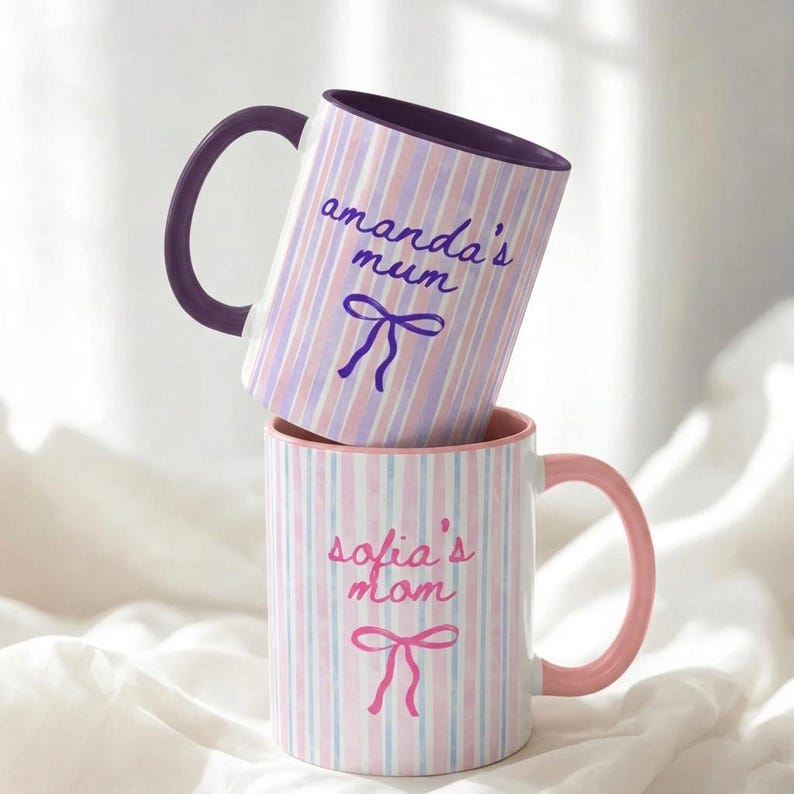 May include: Two ceramic mugs stacked on top of each other. The top mug has a purple handle and the text "amanda's mum" in cursive, with a bow illustration. The bottom mug has a pink handle and the text "sofia's mom" with a bow illustration.
