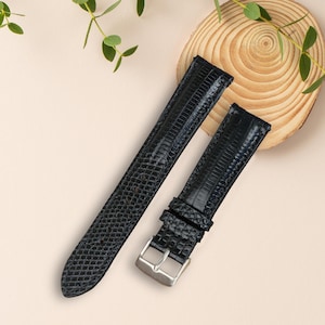 May include: Two black watch straps with a textured, reptile-like pattern. One strap is longer, and the other has a silver-colored buckle. The straps are positioned on a light beige surface with green sprigs and a wooden slice in the background.