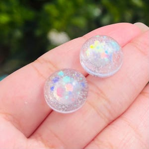 May include: A pair of clear resin stud earrings with iridescent glitter embedded inside. The glitter is in shades of pink, blue, and green.