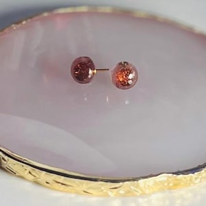 Può includere: Un paio di piccoli orecchini rotondi di colore rosso-marrone con un perno dorato.