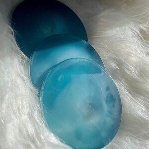 May include: Three round, translucent coasters in shades of blue, arranged on a fluffy white surface. The coasters have a marbled appearance, with lighter blue centers and darker blue edges.