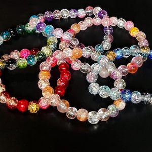 May include: Several colorful beaded bracelets in shades of red, orange, pink, purple, blue, green, and clear. The beads have a crackled appearance and are strung on elastic. The bracelets are displayed on a black surface.