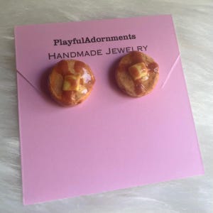 May include: A pair of stud earrings shaped like miniature pancakes with a pat of butter and syrup, attached to a light pink display card printed with the text PlayfulAdornments HANDMADE JEWELRY. The pancakes are golden brown and glossy.