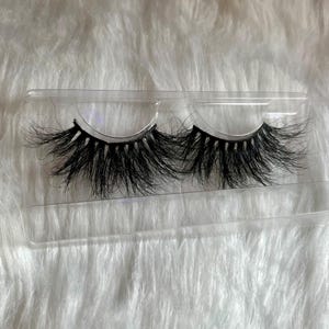 Puede incluir: Un par de pestañas postizas negras y dramáticas en un estuche de plástico transparente. Las pestañas son largas, gruesas y esponjosas, con un diseño en capas para mayor volumen. El estuche está sobre una superficie blanca texturizada.