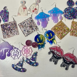 Puede incluir: Una colección de pendientes hechos a mano en varias formas y colores. Los diseños incluyen cuadrados llenos de purpurina, formas de champiñones, sombreros de bruja y labios goteando. Los pendientes son en tonos de azul, morado, rosa y dorado.