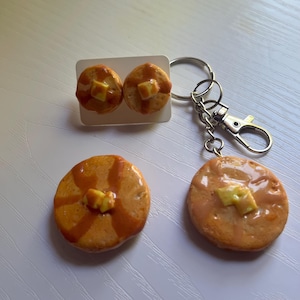 Op de afbeelding: Een set van twee ronde pannenkoekvormige oorknopjes op een witte kaart, samen met een ronde pannenkoekvormige sleutelhanger met een zilveren metalen ring en karabijnhaak. Elke pannenkoek heeft een glanzende siroopafwerking en een klein klontje boter.