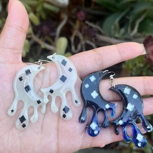 May include: A pair of handmade earrings in a crescent moon shape with a dripping design. One pair is a translucent white with black square accents, and the other is dark blue with white square accents.