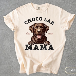 May include: A cream-colored t-shirt with a brown chocolate lab peeking out from the bottom. The shirt has black text that reads "Choco Lab Est. 2025 Mama".