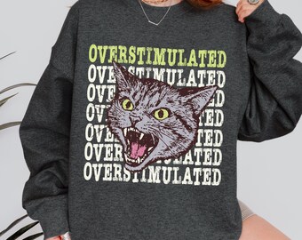 May include: Dark heather gray sweatshirt with the text "OVERSTIMULATED" stacked vertically. A distressed illustration of a gray cat with yellow eyes and an open mouth is in the center. The words "DARK HEATHER" are at the bottom.