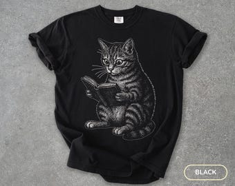 Könnte beinhalten: Schwarzes T-Shirt mit einer detaillierten Illustration einer Katze, die ein Buch liest. Die Katze ist in sitzender Position dargestellt, mit weit aufgerissenen Augen, die auf das Buch gerichtet sind. Das Design ist in Schwarzweiß gehalten und erzeugt eine klassische, Vintage-Ästhetik.