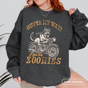 Peut inclure: Sweat-shirt gris foncé avec un dessin d'un chien à moto, portant un casque. Le texte "OUTTA MY WAY! I got the ZOOMIES" est en orange. Sweat-shirt de style décontracté.