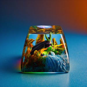 Puede incluir: Una tecla personalizada con un personaje verde y negro con capa, dentro de un bloque de resina transparente. La tecla tiene un diseño detallado con detalles amarillos y naranjas, sobre un fondo azul.