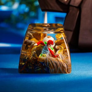 Puede incluir: Una tecla de resina transparente y rectangular con una figura en miniatura detallada de un personaje con ropa roja y verde. La figura se encuentra sobre un fondo de texturas marrones y doradas, con un fondo azul.