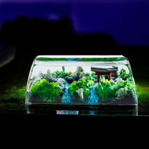 May include: A clear, rectangular resin keycap featuring a miniature landscape. The scene includes green foliage, gray rock formations, and a small red structure, with blue water features. The keycap is designed to fit on a mechanical keyboard.