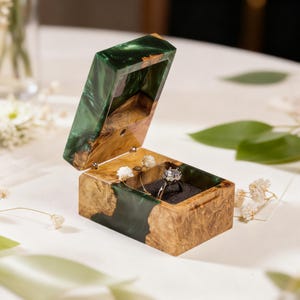 Könnte beinhalten: Geöffnete Holz-Ringbox mit grün-braunem Harz-Außen. Im Inneren liegt ein Diamantring auf einem schwarzen Kissen. Die Box ist mit kleinen weißen Blumen dekoriert, passend für eine Hochzeit oder Verlobung.