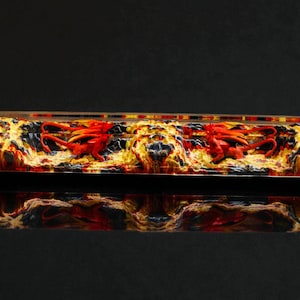 Puede incluir: Una tecla rectangular decorativa con un diseño de fuego. La tecla presenta una mezcla de colores rojo, amarillo y negro, con una figura de dragón en el centro. La tecla se refleja en una superficie negra.