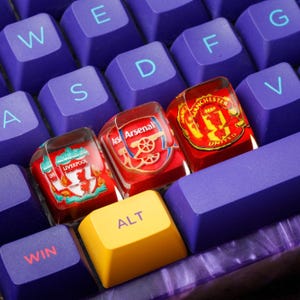 May include: A close-up of a mechanical keyboard with purple keycaps and custom keycaps featuring football team logos. The ALT key is yellow. The keycaps display the logos of Liverpool, Arsenal, and Manchester United.