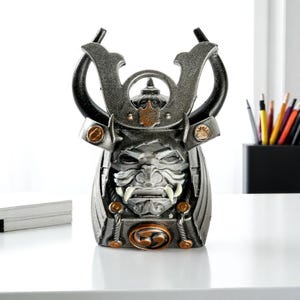 Op de afbeelding: Gedetailleerde, metalen samoeraihelmsculptuur met een fel gezicht, prominente hoektanden en een decoratief embleem. De helm heeft een donkergrijze en bronzen kleurstelling, met gebogen hoorns en sierlijke details. De sculptuur staat op een wit oppervlak.