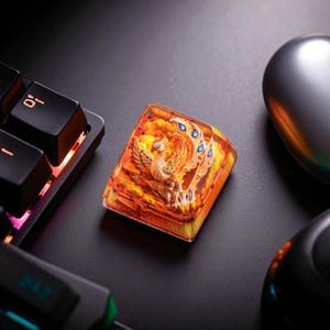 Könnte beinhalten: Eine individuelle Tastaturkappe mit einem Phönix-Design. Die Kappe ist hauptsächlich orange und gelb, mit einem geformten Phönix in der Mitte. Der Phönix hat blaue Akzente und ist vor einem feurigen Hintergrund platziert. Die Kappe liegt auf einer schwarzen Oberfläche.
