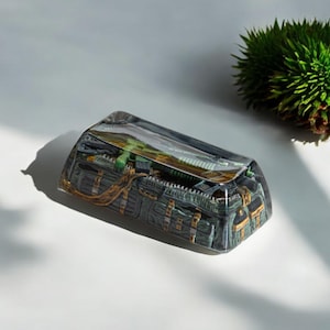 Op de afbeelding: Een rechthoekige, heldere hars keycap met een gedetailleerde miniatuurweergave van een militaire duffeltas. De tas is voornamelijk donkergroen met gouden accenten en een rits. Een groen geweer is zichtbaar in de tas.