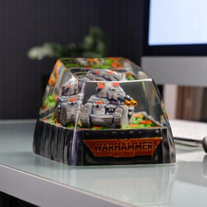 May include: A clear plastic display case with a black base and a silver and orange Warhammer 40,000 tank inside. The case has a black base with the Warhammer 40,000 logo on it.