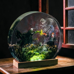 Può includere: Una scultura sferica in resina trasparente raffigurante una scena di battaglia magica con figure in miniatura. La scena è ambientata all'interno di un globo, con una base muschiosa ed effetti di fumo vorticoso. La scultura è montata su una base di legno.