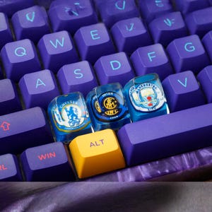 Puede incluir: Primer plano de un teclado mecánico con teclas moradas y una tecla "ALT" amarilla. Tres teclas muestran logotipos de clubes de fútbol: Chelsea, Inter de Milán y Manchester City. El teclado tiene un diseño veteado morado y blanco.