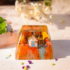 May include: A clear, rectangular resin art piece featuring a miniature landscape. The scene includes a white house with blue window frames, a waterfall, and orange and brown foliage. The resin has a yellow-orange hue.