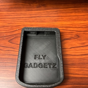 Puede incluir: Un soporte negro rectangular para gadgets con el texto "FLY GADGETZ" en relieve en la superficie. El soporte tiene un borde texturizado y está diseñado para sujetar un pequeño dispositivo electrónico.