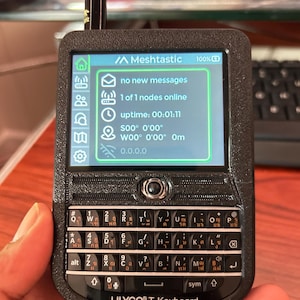Op de afbeelding: Zwart handheld apparaat met een scherm met "Meshtastic" en andere gegevens. Het heeft een fysiek toetsenbord met Thaise tekens en de tekst "LILYGO T-Keyboard". Een antenne steekt uit de bovenkant.