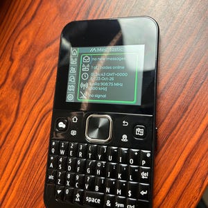 May include: A black handheld Meshtastic communication device with a QWERTY keyboard and a digital screen displaying no new messages, 1 of 1 nodes online, the date Fri 23-Oct-26, and LoRa 908.75 MHz 500 kHz frequency information.