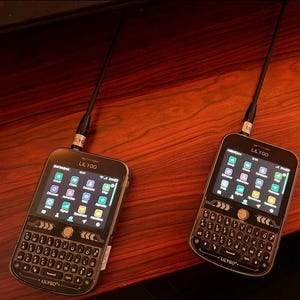 Op de afbeelding: Twee zwarte LILYGO draagbare communicatieapparaten met QWERTY-toetsenborden en kleuren-touchscreens. Elk apparaat is voorzien van een antenne aan de bovenzijde, een centrale navigatieknop en een digitaal scherm met diverse app-pictogrammen.