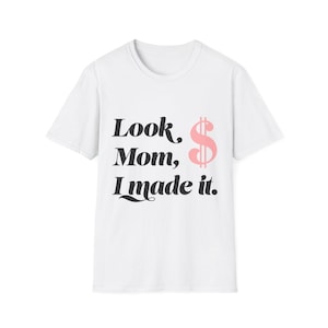 May include: White t-shirt with the phrase "Look Mom, I made it." in black text, accompanied by a pink dollar sign. The shirt has a crew neck and short sleeves, suitable for casual wear.