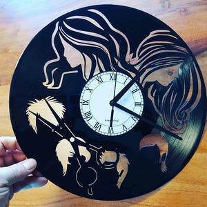 Op de afbeelding: Een zwarte vinyl platenspeler klok met een witte wijzerplaat en zwarte wijzers. De wijzerplaat heeft Romeinse cijfers en de tekst "Kreativ Vinyl". De klok is versierd met een silhouet van twee vrouwenhoofden en een schaar.