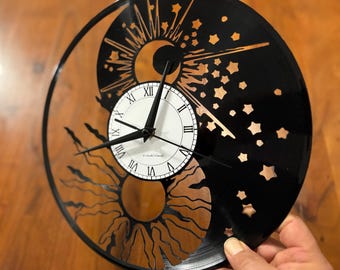 Puede incluir: Un reloj de vinilo negro con una esfera blanca y números romanos. La esfera está rodeada por un diseño de un sol y una luna con estrellas. El reloj está hecho de un disco de vinilo reutilizado.