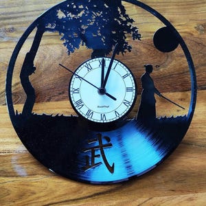 Puede incluir: Un reloj de vinilo negro con una esfera blanca y agujas negras. El reloj está decorado con una silueta de un árbol, una persona sosteniendo una espada y la palabra japonesa "Bushido" en letras doradas.
