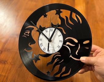 Puede incluir: Reloj de vinilo negro con un diseño de sol y luna. La esfera del reloj es blanca con marcadores de hora romanos negros y manecillas negras.