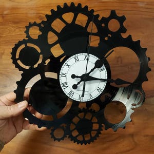 May include: A black wall clock in a gear design, with a white clock face and black Roman numerals. The clock hands are black. The clock is held against a wooden background.