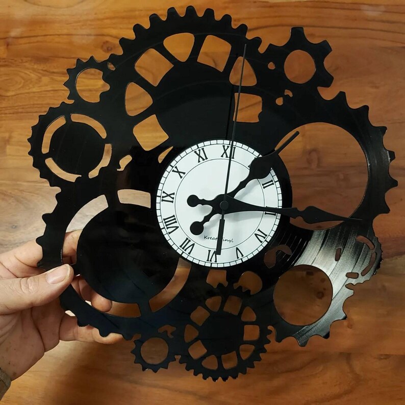 May include: A black wall clock in a gear design, with a white clock face and black Roman numerals. The clock hands are black. The clock is held against a wooden background.