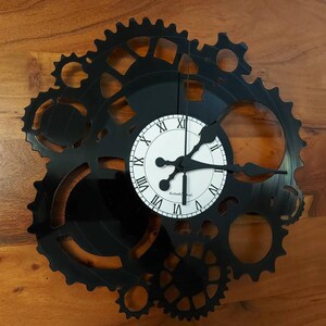 May include: Black wall clock with a gear design. The clock face is white with black Roman numerals and hands. The clock is made from a repurposed vinyl record, offering a unique, industrial aesthetic.
