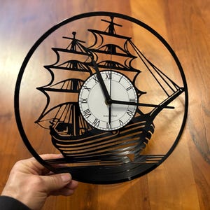 Könnte beinhalten: Schwarze Schallplattenuhr mit einer Silhouette eines Segelschiffs. Das Zifferblatt der Uhr ist weiß mit schwarzen römischen Ziffern und schwarzen Zeigern.