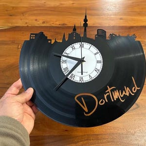 Puede incluir: Un reloj de vinilo negro con una silueta de horizonte urbano. La esfera del reloj es blanca con números romanos y manecillas negras. La palabra "Dortmund" está escrita en color cobre en el disco.