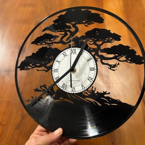 Op de afbeelding: Zwarte vinylplatenklok met een uitgesneden bonsai-boomontwerp. De klok heeft Romeinse cijfers en zwarte wijzers. Een unieke decoratieve klok.