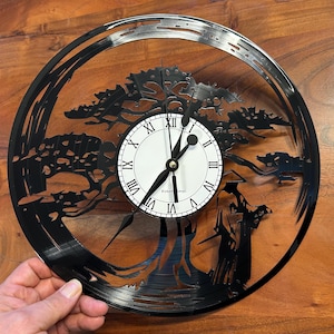 Op de afbeelding: Zwarte vinyl platen klok met een silhouet van een boom en een persoon in een gewaad. De wijzerplaat van de klok is wit met zwarte Romeinse cijfers en zwarte wijzers. De klok heeft een diameter van 30,5 cm.