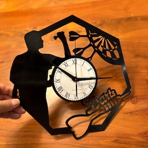 May include: A black vinyl record clock with a silhouette of a person throwing darts at a dartboard. The clock face is white with black Roman numeral markings. The clock is shaped like a hexagon and has the word "Darts" written on it.