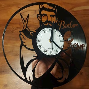Op de afbeelding: Zwarte vinyl platen klok met een silhouet van een barbier die een schaar en een scheermes vasthoudt. De wijzerplaat van de klok is wit met zwarte Romeinse cijfers en de woorden "Kreatvinyl" onderaan. De woorden "Barber Shop" staan op de plaat.